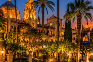Panoramic evening illustration of the illuminated historic Mission Inn Hotel, Spanish Revival architecture, and downtown streets in Riverside, California.